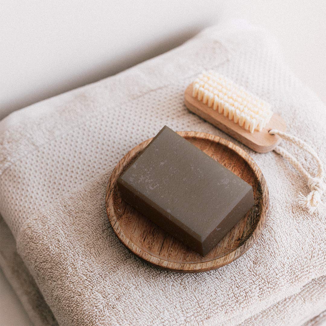 An overhead view of &Better's body scrub bar placed on a wooden soap dish, next to a soft cotton towel, set against a minimalist background, highlighting the simplicity and natural appeal of the product