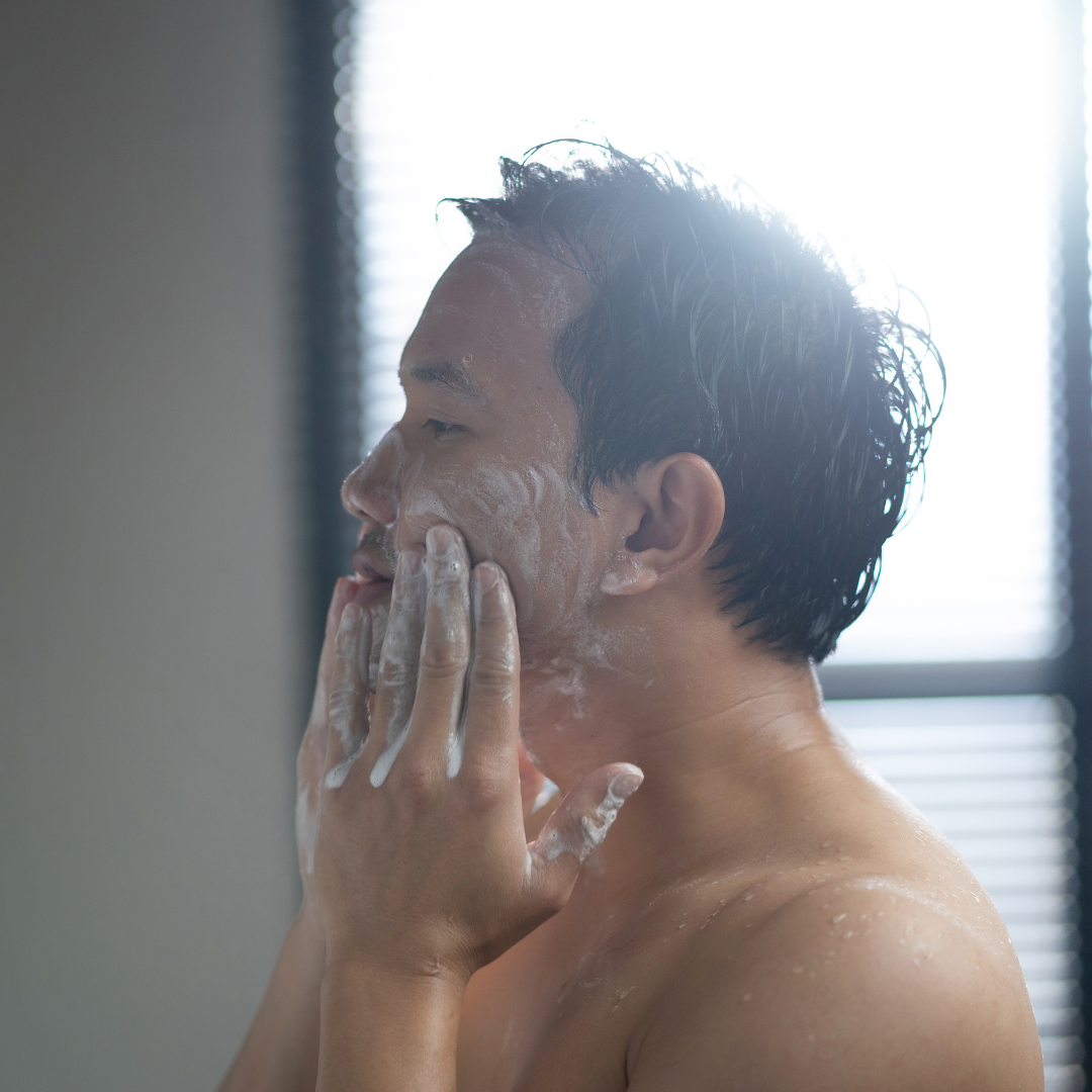 A young man with a towel wrapped around his waist applying face cream with his fingers after taking a shower.
