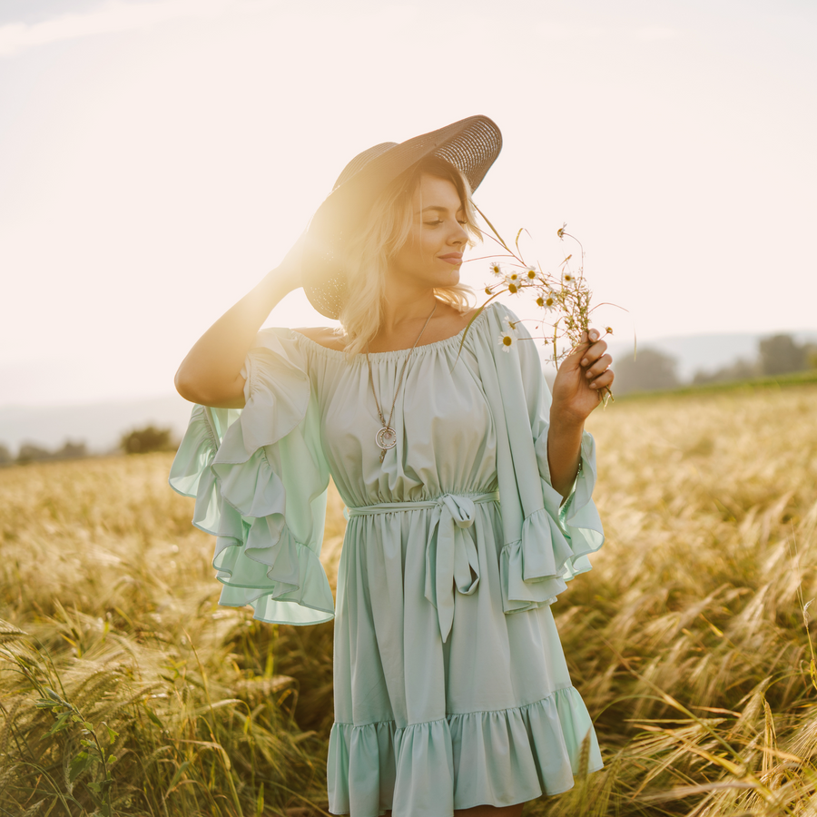 A beautiful lady with a nice cotton dress and hat smelling flowers on a green field on sunset